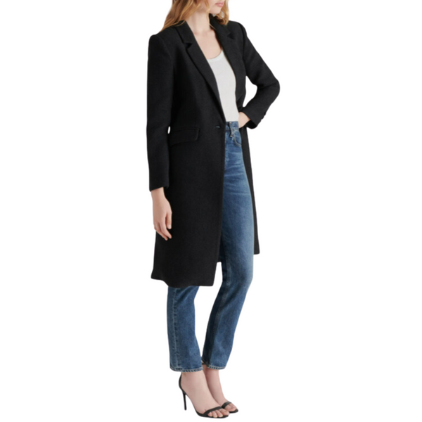 Woman in a black coat, white top, blue jeans, and black heels.