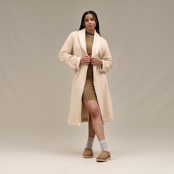 Person in a beige coat, olive dress, striped socks, and tan shoes standing against a neutral background.