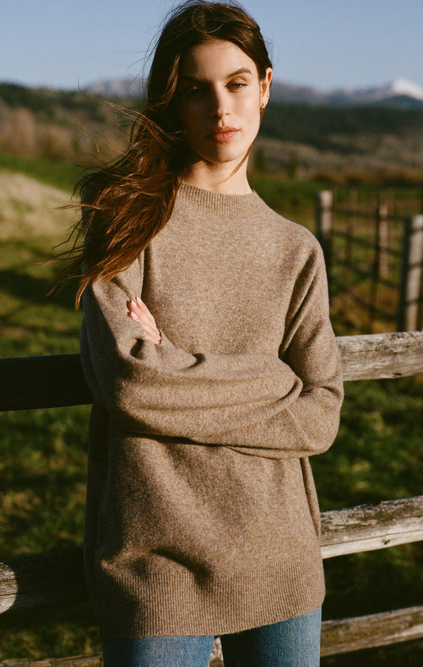 Woman in a brown sweater and jeans standing by a wooden fence outdoors.